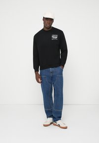 Junger Mann mit weißer Mütze, schwarzem Sweatshirt mit Auto-Grafik, blauen Jeans mit sichtbaren Nähten und weißen Turnschuhen, der vor einem einfarbigen Hintergrund steht.