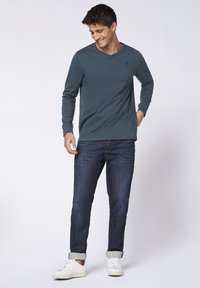Langärmliges, türkisfarbenes Henley-Shirt mit Knöpfen, kombiniert mit dunklen Jeans und weißen Sneakers. Das Outfit zeichnet sich durch ein lässiges, modernes Design aus.
