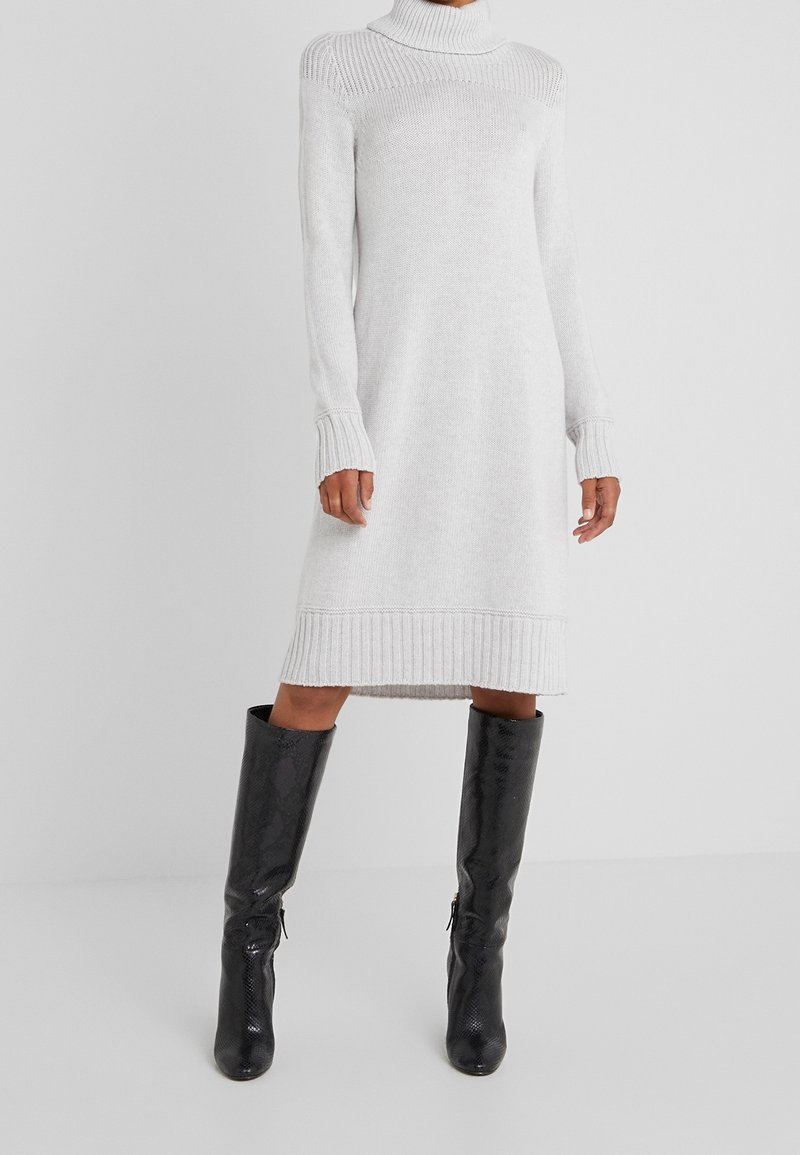 Personne portant une robe midi en maille blanche à manches longues avec un col roulé et des bottes noires texturées montant jusqu’aux genoux, debout sur un fond uni.