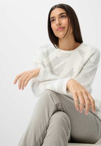 Weißer und hellgrauer Strickpullover mit zigzagförmigem Muster, kombiniert mit hellgrauen Hosen. Merkmale sind die weiche Textur und ein zartes Ring.