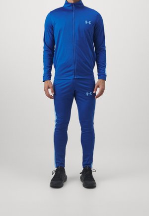 Personne portant un survêtement bleu Under Armour avec une veste zippée et un pantalon assorti, assorti à des chaussures de sport noires.