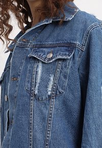 Farkkutakki keskipunaisena, jossa on kulutetut yksityiskohdat, nappikiinnitys, yksi rintatasku ja viistot kaulukset. Teksturoitu kangas ja rento malli.