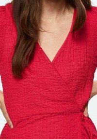 Czerwona, teksturowana sukienka wrap z dekoltem w kształcie V i wiązaniem w talii; tkanina ma marszczoną powierzchnię, co tworzy dynamiczny efekt wizualny.