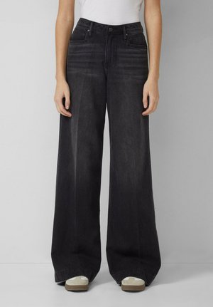 Femme portant un jean noir taille haute à jambes larges avec détails délavés, assorti de baskets blanches et d'un haut blanc rentré, debout contre un mur uni.