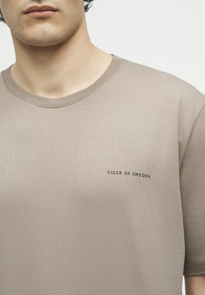 Κοντινό πλάνο ενός ατόμου που φοράει μπεζ T-shirt με την επιγραφή «TIGER OF SWEDEN» στο αριστερό στήθος.