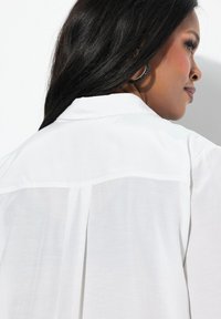 Camicia bianca con bottoni, caratterizzata da un design a pieghe sul retro e un colletto classico. Tessuto morbido e leggero con una superficie liscia.