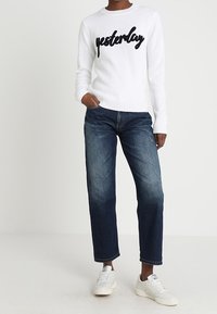 Biały dziergany sweter z napisem „yesterday” w czarnym, kursywnym stylu, zestawiony z ciemnoniebieskimi, luźnymi dżinsami i białymi trampkami. Prosty design, casualowy wygląd.