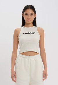 Lichtbeige cropped tanktop van ribstof met een ronde halslijn en zwart "lowlights" logo op de voorkant, gecombineerd met witte joggingbroeken.