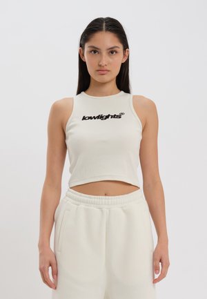 Haut de tank top court beige clair en tissu côtelé avec un col rond et le logo "lowlights" noir sur le devant, associé à un pantalon de jogging blanc.