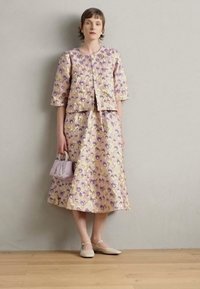 Completo due pezzi in jacquard floreale lilla, composto da una giacca corta e una lunga gonna. Gli accessori includono una piccola borsa rosa e scarpe beige.