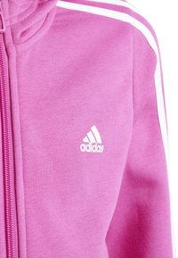 Sudadera de cremallera de color rosa claro, hecha de tela suave, con detalles de franjas blancas y un pequeño logotipo blanco de Adidas en el pecho.