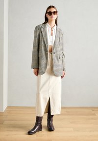 Gepatchter Blazer in Schwarz- und Cremetönen, weißes Hemd, beiger Midirock mit Schlitzen vorne, braune Knöchelstiefel und dunkle Sonnenbrille.