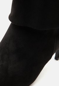 Κοντινή λήψη μαύρης σουέντ μποτάκια με ψηλό τακούνι, που δείχνει τη μυτερή μύτη και τη λεπτομέρεια με διπλωμένη μανσέτα.