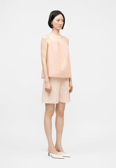 Γυναίκα που στέκεται φορώντας ροζ παστέλ και λευκό ριγέ αμάνικο τοπ, ταιριαστά φαρδιά σορτς και κρεμ παπούτσια με μυτερή μύτη σε απλό φόντο.