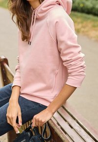 Felpa rosa chiaro con cappuccio e cordoncino, dotata di polsini a costine, tessuto morbido e tasca a marsupio. Abbinata a jeans scuri e una borsa.
