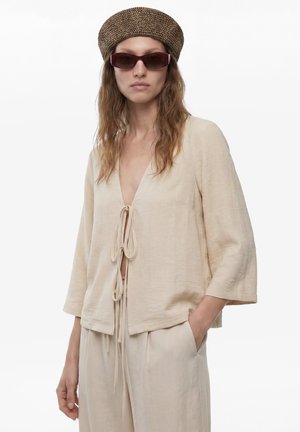 Femme portant un haut ample beige avec liens devant, pantalon assorti, chapeau tressé marron et lunettes de soleil foncées, posant avec une main dans la poche.