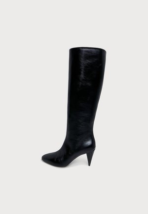 Botte noire en cuir montant jusqu’au genou, à bout pointu et à talon haut et fin, sur un fond uni clair.