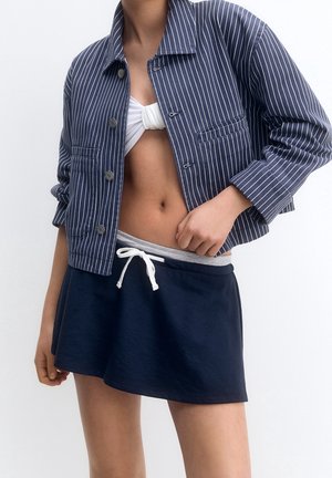 Personne portant une veste bleu marine à fines rayures, un crop top blanc noué et une jupe bleu marine avec un cordon blanc, debout devant un fond blanc.