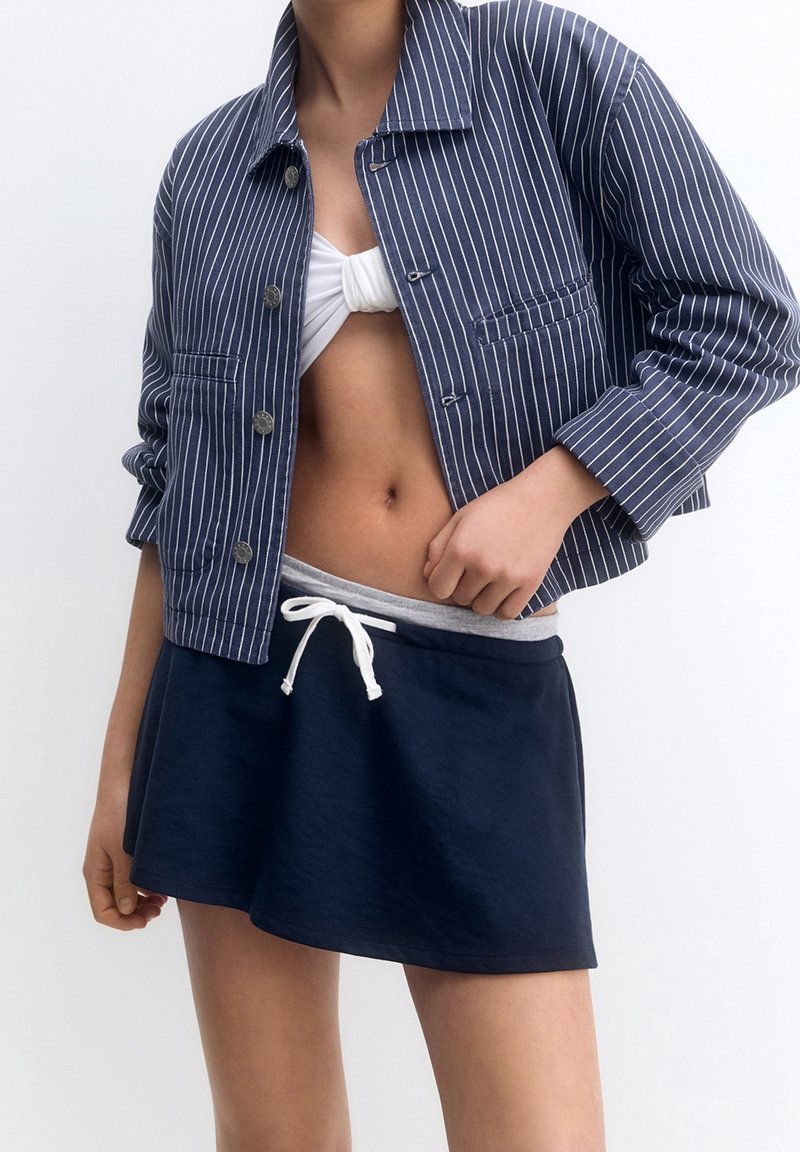 Personne portant une veste bleu marine à fines rayures, un crop top blanc noué et une jupe bleu marine avec un cordon blanc, debout devant un fond blanc.