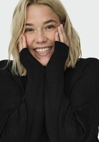 Maglione nero a maglia con maniche lunghe, dotato di polsini a coste. Testura morbida e materiale leggero, indossato da una persona con un sorriso luminoso.