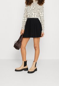 Svart plisserad kjol, mönstrad långärmad topp i crème, beiga ankelstövlar med svart elastik och en brun satinklänka.