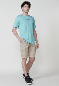 Hellblaues T-Shirt mit Logo, kombiniert mit beigen Cargo-Shorts mit mehreren Taschen. Schwarze Sportschuhe vervollständigen das Outfit.