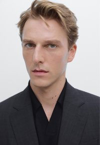 Porträtt av en person med ljust brunt hår och gröna ögon, klädd i en svart skjorta med krage under en mörk mönstrad blazer mot en vit bakgrund.