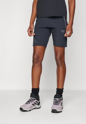 Atletisk person iført sorte shorts, sorte sokker og lyse lilla og sorte løbesko, stående mod en ensfarvet hvid baggrund.