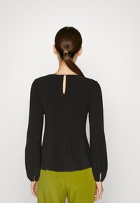 Top nero a maniche lunghe con costine, caratterizzato da un dettaglio a oblò sulla parte posteriore del collo, con texture arricciata e polsini aderenti. Abbinato a pantaloni verde.