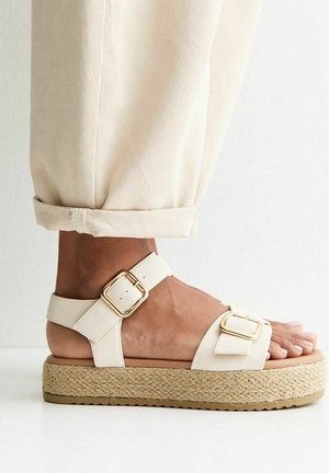 Pie con sandalia beige de plataforma con suela de yute trenzado y tiras con hebilla dorada, combinado con pantalones beige claro remangados.