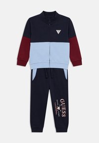 Σκούρο μπλε φόρμα με λεπτομέρειες σε ανοιχτό μπλε και μπορντό. Περιλαμβάνει μπουφάν με φερμουάρ και παντελόνι με τσέπες και τυπωμένο λογότυπο "GUESS".
