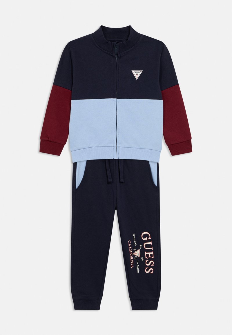 Σκούρο μπλε φόρμα με λεπτομέρειες σε ανοιχτό μπλε και μπορντό. Περιλαμβάνει μπουφάν με φερμουάρ και παντελόνι με τσέπες και τυπωμένο λογότυπο "GUESS".