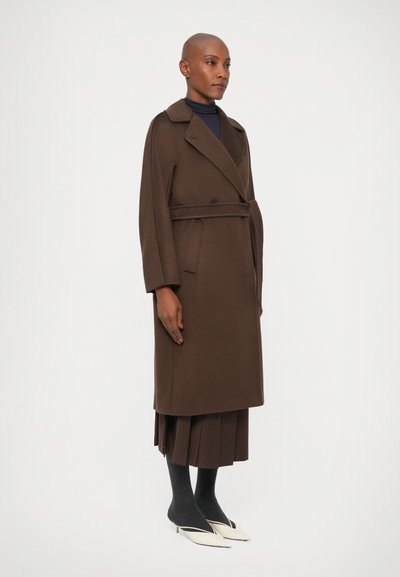 Brun uld trenchcoat med bælte i taljen, brede revers og sidelommer, lagd over en plisseret brun nederdel og mørke strømpebukser. Hvide spidse sko.