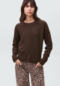 Femeie îmbrăcată cu un pulover brown cu guler rotund și detalii cu nasturi, purtând pantaloni cu imprimeu leopard și talie înaltă, stând cu mâinile în buzunare.