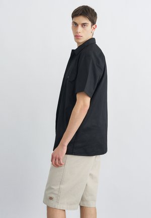 Giovane uomo in piedi di profilo che indossa una maglietta nera a maniche corte e pantaloncini beige fino al ginocchio su uno sfondo bianco uniforme.