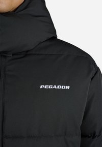 Črna puhovka z visokim ovratnikom, okrašena z belim vezenim logotipom "PEGADOR". Gladka tekstura, prešite zasnovo in sprednja zadrga.
