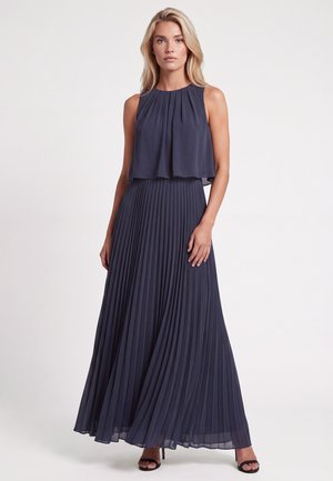 Frau trägt ein ärmelloses, marineblaues plissiertes Maxikleid mit einem fließenden, geschichteten Top und steht vor einem schlichten hellen Hintergrund.
