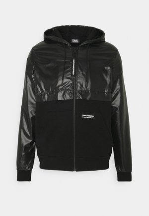 Haut de sweat à capuche zippé noir avec un dessus brillant et un bas mat, doté d'une poche kangourou et d'une capuche à cordon de serrage, réalisé en un matériau lisse et doux.