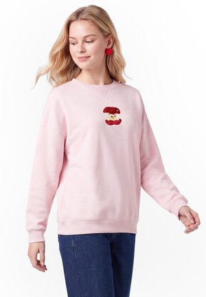Giovane donna che indossa un maglione rosa con una toppa a fungo rossa e orecchini rossi a forma di cuore, abbinati a jeans blu, in piedi su sfondo bianco.