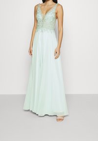 Dlouhá mátová zelená večerní šaty s hlubokým výstřihem ve tvaru V, zdobeným živůtkem a tenkými ramínky. Tkanina má pěknou, lehkou strukturu.