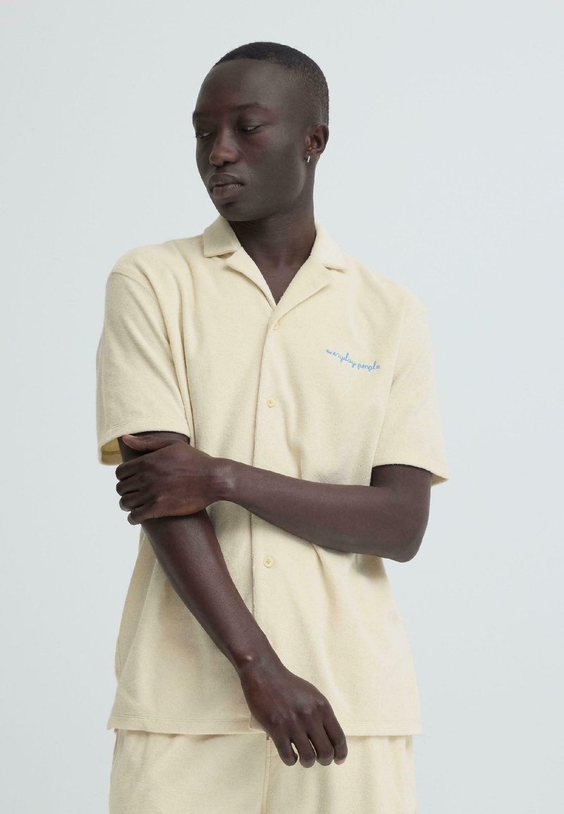 Personne à la peau foncée portant une chemise beige à manches courtes et un short, regardant vers la gauche, avec un bras croisé tenant l'avant-bras opposé.