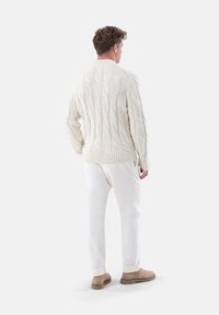Kuscheliger Pullover aus dickem Material mit Kabelstrickmuster, kombiniert mit weißen Hosen und beigen Schuhen. Der Pullover hat einen gerippten Saum und Bündchen.
