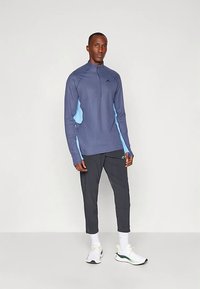 Muškarac odjeven u tamnoplavu i svijetloplavu dugorukavu majicu sa zipom, crne sportske hlače, bijele čarape i bijele tenisice stoji uz jednostavnu pozadinu.