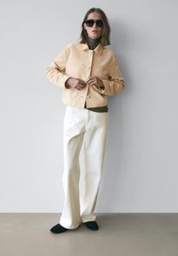 Beige ruskindjakke med knapper, kombineret med en mørkegrøn top og cremefarvede bukser med vide ben. Båret med sorte slip-on sko og solbriller.