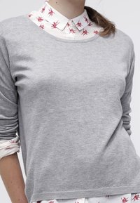 Grauer Strickpullover mit langen Ärmeln und rundem Ausschnitt, über einem weißen Hemd mit Kragen, das rote Blättermuster zeigt.