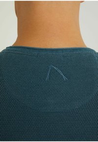 Close-up of a person's neck and upper back wearing a textured, dark teal sweater with a small stitched logo near the neckline.