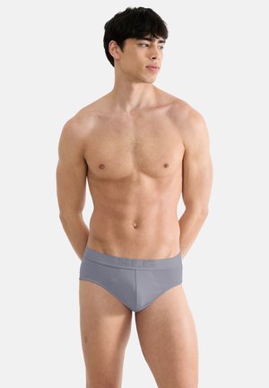 Man zonder shirt met zwart haar, dragend een grijze slip, staand tegen een effen lichte achtergrond, kijkt naar de zijkant met handen achter de rug.