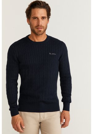 Mörkblå kabelstickad tröja med ribbade ärmslut och nederkant, med en subtil broderad logotyp nära halsringningen. Buren över ljusa byxor.