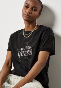 Camiseta de algodón negra con el texto "RODEO QUEEN" en blanco, adornada con tachuelas doradas, mangas cortas y puños enrollados.
