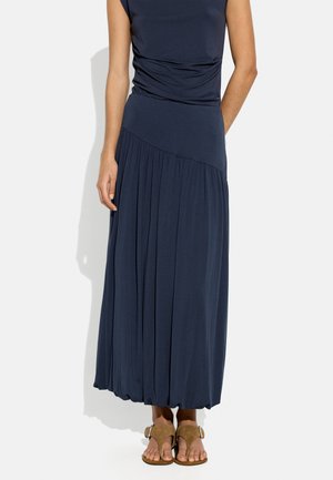 Kobieta nosząca granatowy bezrękawnik i plisowaną spódnicę maxi z brązowymi sandałami z odkrytymi palcami, stojąca na białym tle.